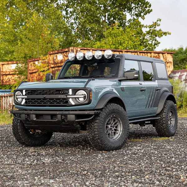 Customized off-road SUV with roof lights parked on gravel, surrounded by trees and rusty containers.