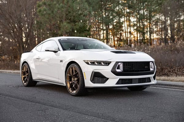 White sports car with sleek design and bronze wheels parked on a road, surrounded by trees.