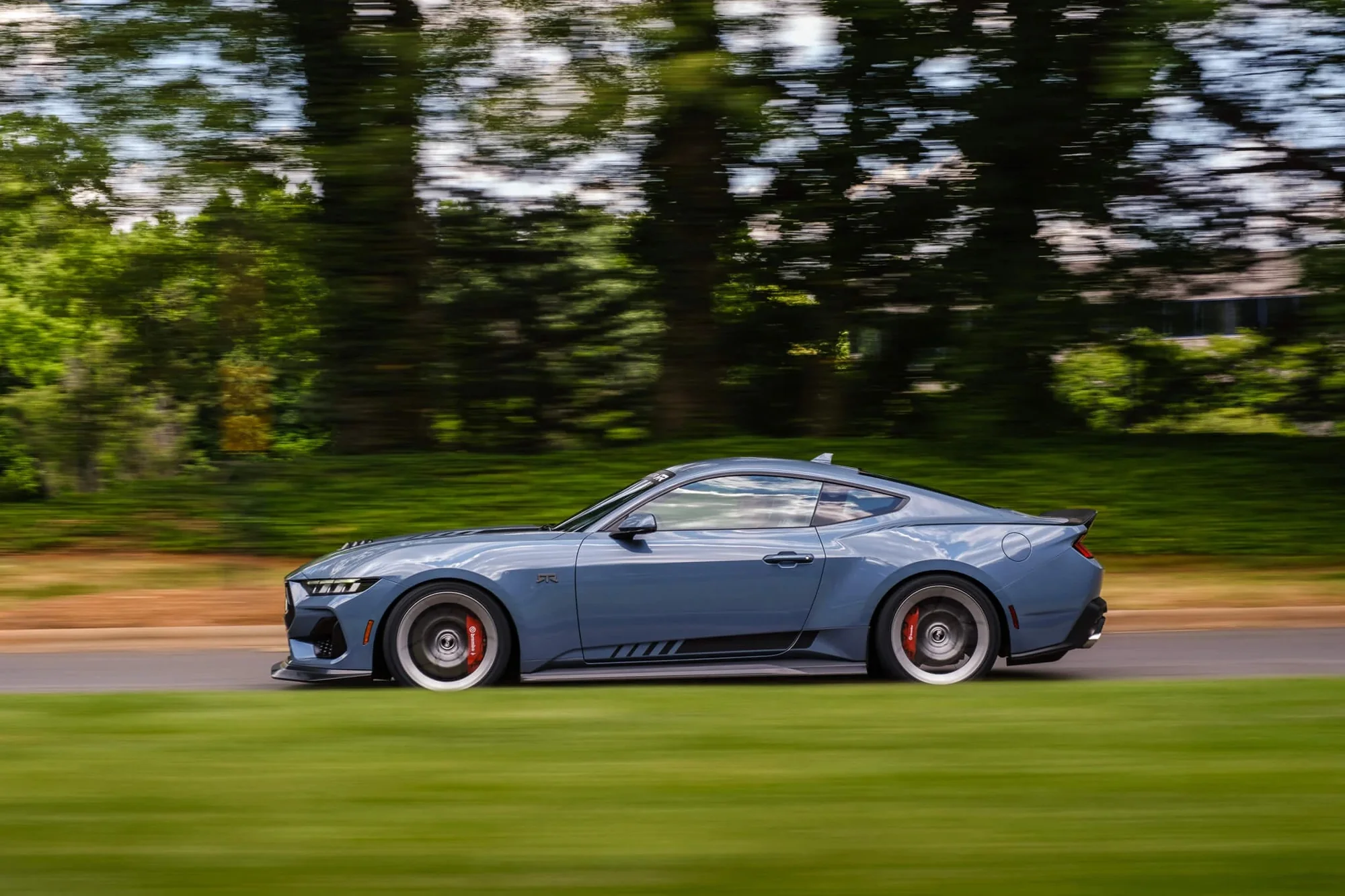 A sleek blue sports car in motion, showcasing its aerodynamic design and sporty features on a tree-lined road. The image captures the vehicle’s aggressive stance and distinctive wheels, emphasizing speed and performance.